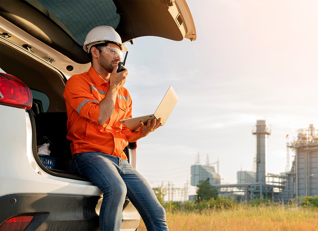 Insurance by Industry - Male Industrial Worker Wearing Safety Uniform Wile Using a Walkie-Talkie and Laptop and Working on an Inspection in a Car by a Power Plant